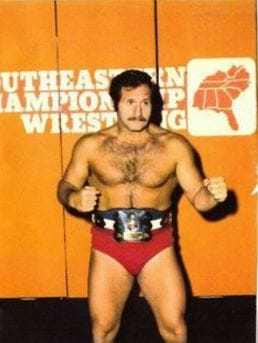 The Great Malenko wearing a wrestling belt posing with his fists up in front of the Southeastern Championship Wrestling Background