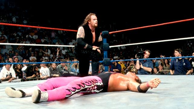 The Undertaker on one knee beside Bret Hart as he lay flat on his back