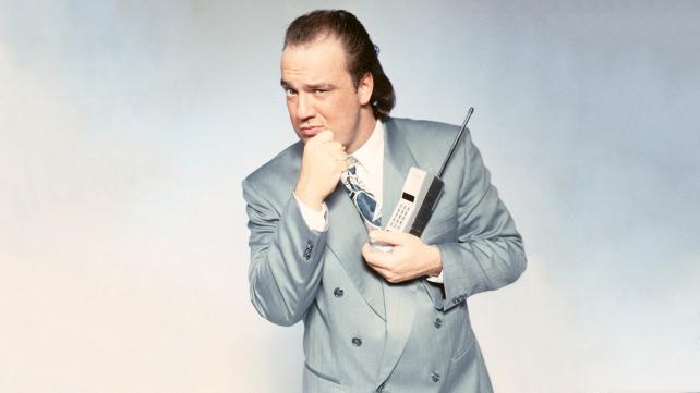 Paul Heyman in a grey suit holding a early 1980's cell phone with his hand up to his chin