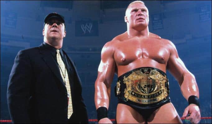 Paul Heyman in a black suit and baseball cap standing next to Brock Lesnar wearing a title belt
