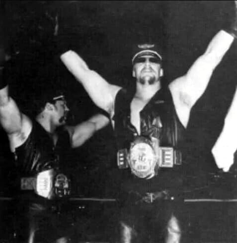 The Road Warriors in all black leather in the ring with title belts on and arms in the air working the crowd