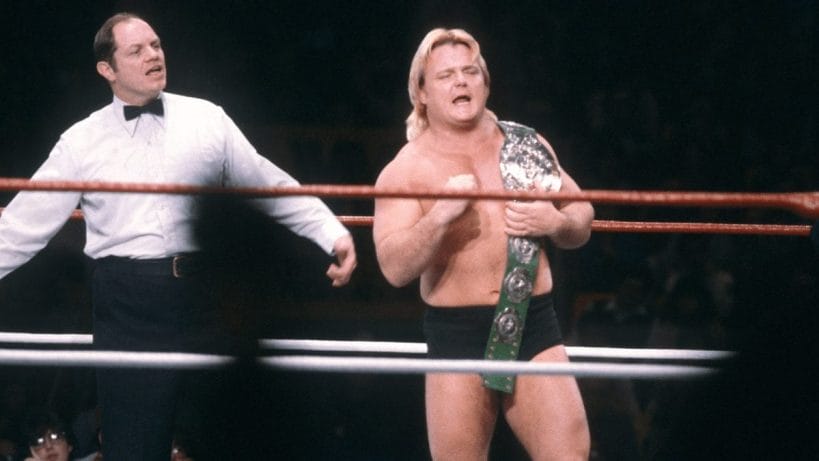 Greg Valentine in a dim-lit wrestling ring holding his green leather Intercontinental Championship belt with a referee behind him
